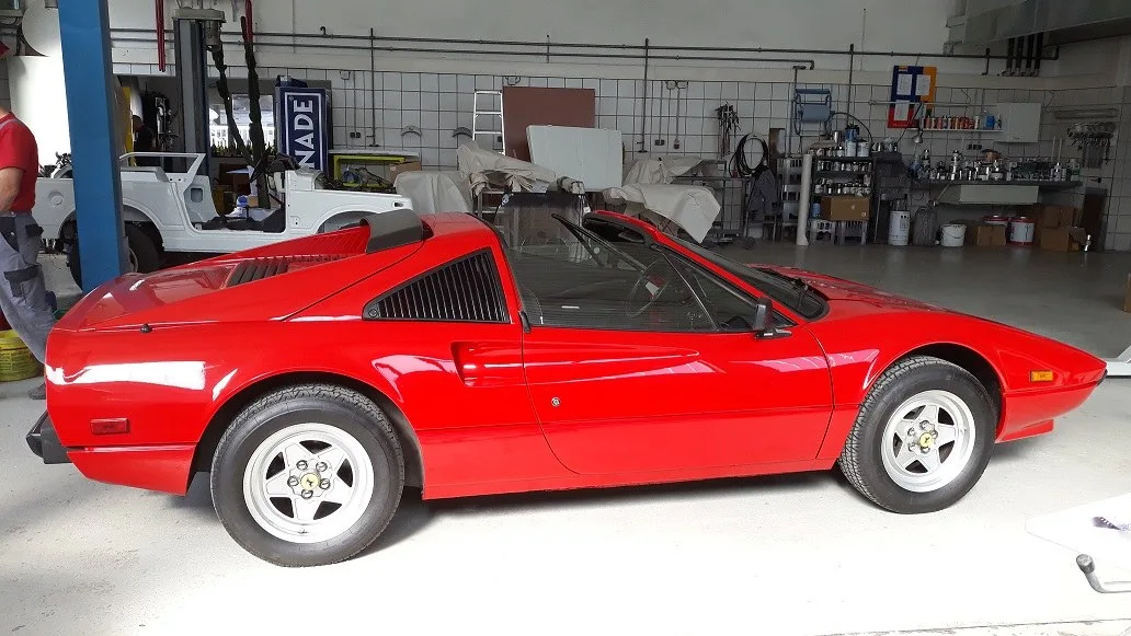 Roter Ferrari 308 GTS in der Werkstatt, dahinter zerlegte Karosserieteile und die Lackierbank