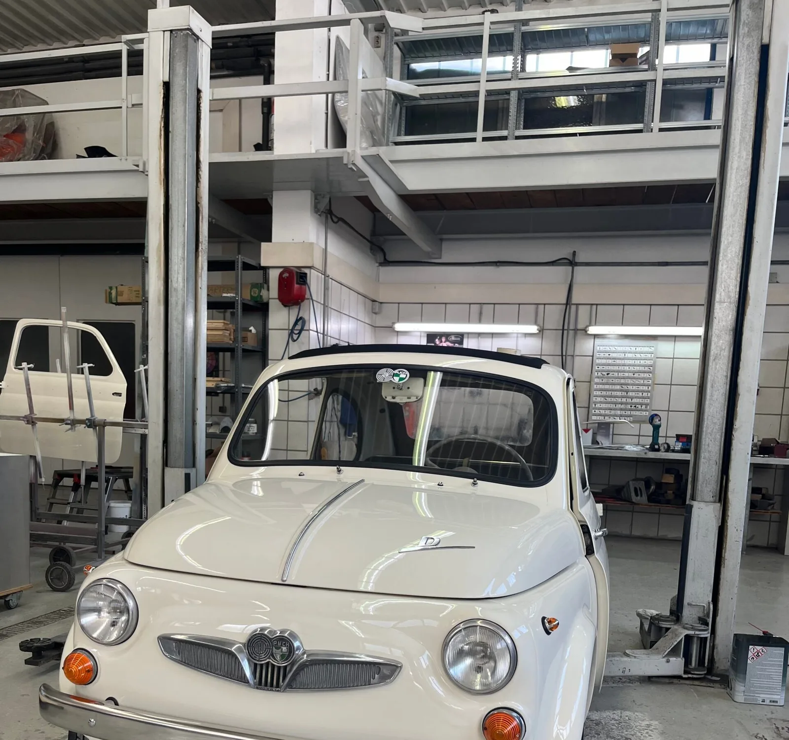 Weißer Steyr-Puch 500, frisch lackiert, zwischen den Hebebühnen der Werkstatt, links eine lackierte Fahrzeugtür auf dem Ständer
